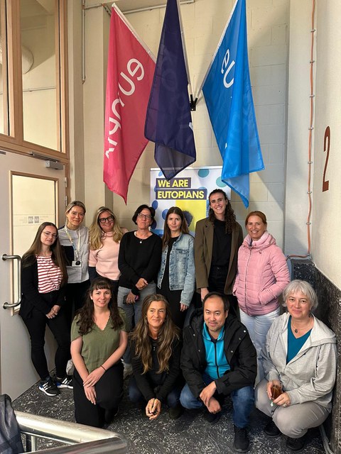 Eine Gruppe von etwa zwölf Personen steht und sitzt für ein Gruppenfoto in einem Innenbereich. Hinter ihnen hängen drei farbige Fahnen und ein Poster mit der Aufschrift „WE ARE EUTOPIANS“. Die Personen lächeln in die Kamera.