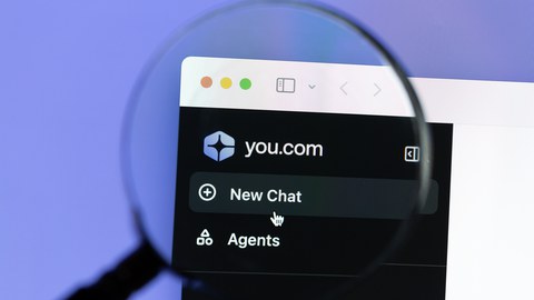 Nahaufnahme eines Computerbildschirms mit geöffneter You.com-Website. Eine Lupe vergrößert einen Teil des Bildschirms, auf dem das Logo von You.com und die Schaltflächen „New Chat“ und „Agents“ zu sehen sind. Der Hintergrund ist lila.