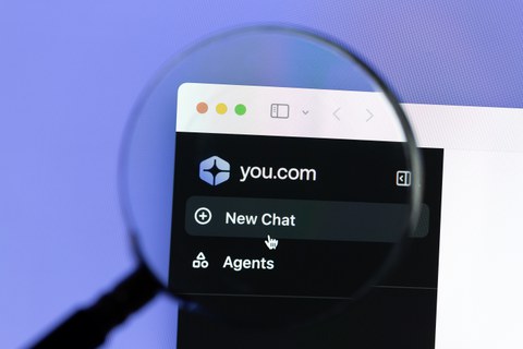 Nahaufnahme eines Computerbildschirms mit geöffneter You.com-Website. Eine Lupe vergrößert einen Teil des Bildschirms, auf dem das Logo von You.com und die Schaltflächen „New Chat“ und „Agents“ zu sehen sind. Der Hintergrund ist lila.