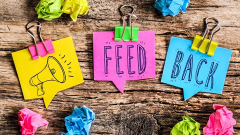 Three colorful sticky notes on a wooden surface display the word "FEEDBACK." The first note, yellow, has a drawing of a megaphone, the second, pink, reads "FEED," and the third, blue, reads "BACK." Around the notes, there are crumpled paper balls in matching colors.