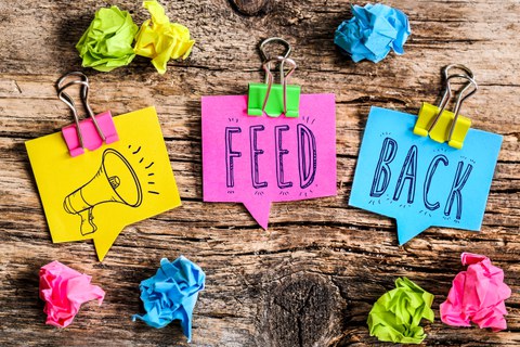 Three colorful sticky notes on a wooden surface display the word "FEEDBACK." The first note, yellow, has a drawing of a megaphone, the second, pink, reads "FEED," and the third, blue, reads "BACK." Around the notes, there are crumpled paper balls in matching colors.