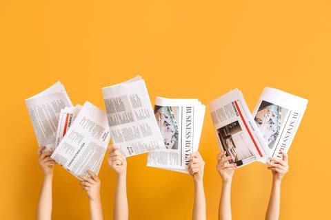 Several people hold open newspapers and magazines in front of their faces. Only their hands and forearms are visible. The publications feature articles about business and finance. The background is solid orange.