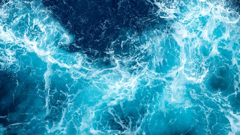 Dynamische Luftaufnahme einer aufgewühlten Meeresoberfläche. Tiefblaue und türkisfarbene Wellen mischen sich mit weißer Gischt, die ein lebendiges und bewegtes Muster auf dem Wasser erzeugt.