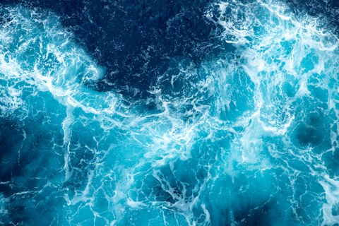 Dynamic aerial view of a turbulent ocean surface. Deep blue and turquoise waves blend with white foam, creating a lively and energetic pattern on the water.