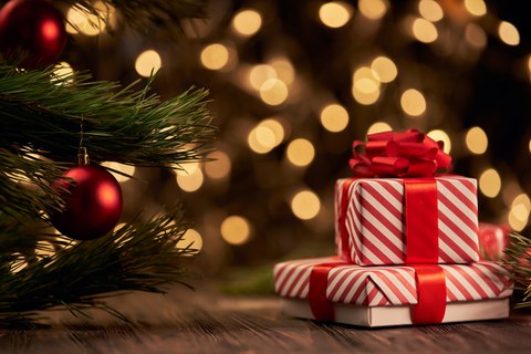 Two red-and-white striped Christmas presents with red ribbons placed next to a decorated pine branch, with warm, blurred lights in the background.
