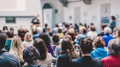 Blick in einen gut besuchten Vortragsraum: Viele Personen sitzen und hören einem unscharf im Hintergrund erkennbaren Vortragenden zu. Die Szene wirkt lebendig und konzentriert.