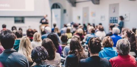 View into a well-attended lecture room: Many people are seated and listening to a presenter, who appears blurred in the background. The scene conveys a lively and focused atmosphere.