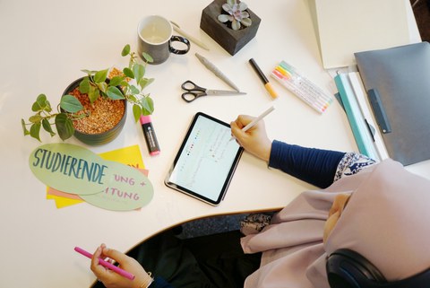 Top view of a desk: A person writes on a tablet with a stylus. On the desk are pens, scissors, markers, note cards including one labeled “Studierende,” a mug, and small potted plants.