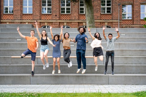 A group of seven young adults jumping in the air together, smiling and cheering. They are standing on gray steps in front of a brick building with large windows. A grassy area is visible in the foreground.