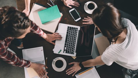 Drei Frauen stützen sich auf einem Tisch ab und schauen von oben auf ihre Laptops und Notizbücher.