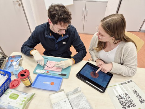 Two students practice a medical suturing technique on a training model. One performs the procedure while the other provides guidance using a digital tutorial on a tablet.