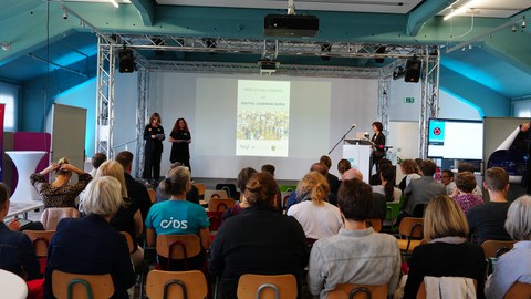 Blick über das Publikum auf die Bühne wo die drei Moderatorinnen sprechen. 