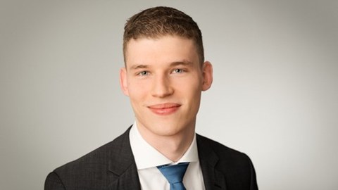 Portrait photo of a young man with short brown hair, fair complexion, and blue eyes. He is wearing a dark suit, a white shirt, and a blue tie. The background is a neutral gray.