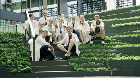 A group of eleven people in white lab coats sits on a green staircase in a modern building. Some are holding scientific objects or cameras. Everyone is smiling and appears cheerful. Glass windows and an architecturally appealing environment are visible in