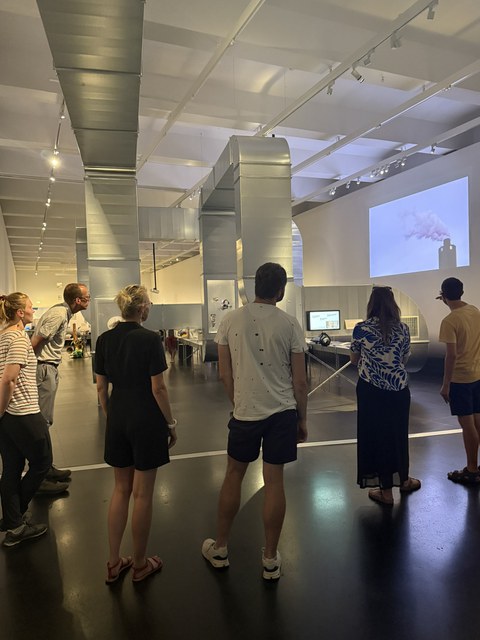 Group of people in an exhibition space with large silver ventilation ducts. A projected image of a chimney emitting smoke is displayed on the wall.