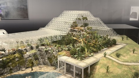 Modell eines großen Gewächshauses oder Biodoms mit pyramidenförmiger Glaskonstruktion über einer künstlich gestalteten Landschaft aus Felsen, Wasser und üppiger Vegetation.