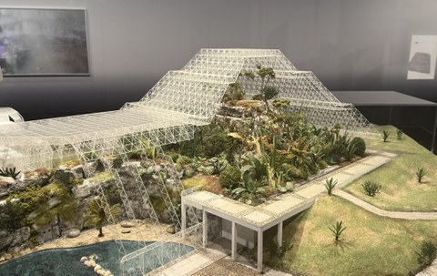 Model of a large greenhouse or biodome with a pyramid-shaped glass structure covering an artificial landscape of rocks, water, and lush vegetation.