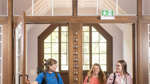 Three students carrying backpacks walk through a large, glasspaned wooden door, laughing, and enter a brightly lit room with a round ceiling light.