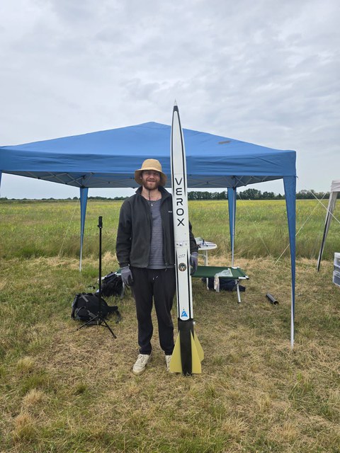 A person wearing a straw hat stands on a meadow in front of a blue canopy, holding a large model rocket labeled “VEROX.” In the background are tables and equipment. The sky is cloudy.