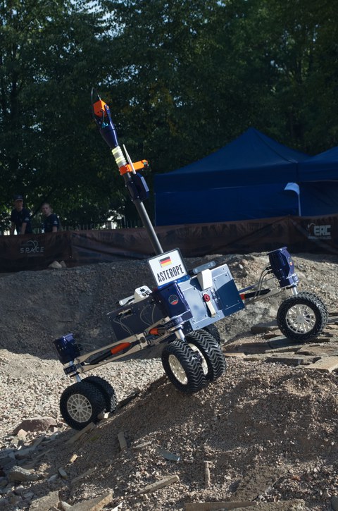 A rover with four large wheels and a robotic arm drives across rough, rocky terrain. A sign reading “ASTEROPE” with the German flag is attached to the vehicle. In the background, a blue tent and trees are visible.
