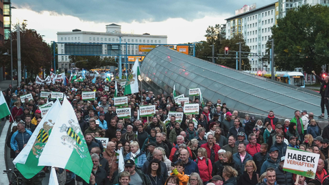  A large crowd at a demonstration in a German city. Many participants hold signs with slogans like “Arrest Habeck,” “Peace with Russia,” and “Out of NATO.” Some carry Saxon flags. Modern buildings and a tram stop are visible in the background.