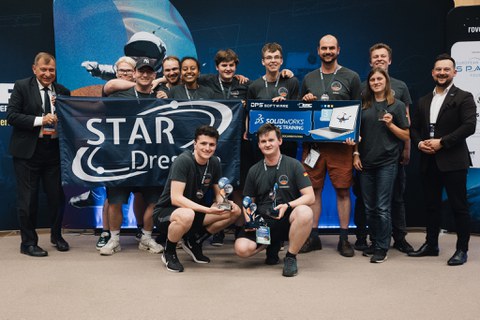 Group of young people in grey team shirts posing at an award ceremony. Two people kneel in front holding trophies, others hold a banner reading “STAR Dresden” and a prize poster. On both sides stand men in suits.