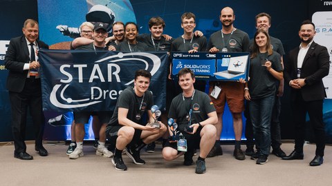 Gruppe junger Menschen in grauen Team-T-Shirts posiert bei einer Siegerehrung. Zwei Personen knien vorn mit Pokalen, andere halten ein Banner mit der Aufschrift „STAR Dresden“ sowie ein Gewinn-Plakat. Links und rechts stehen Anzugträger.