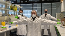 Three young people are standing in a laboratory. They are wearing lab coats and protective masks.