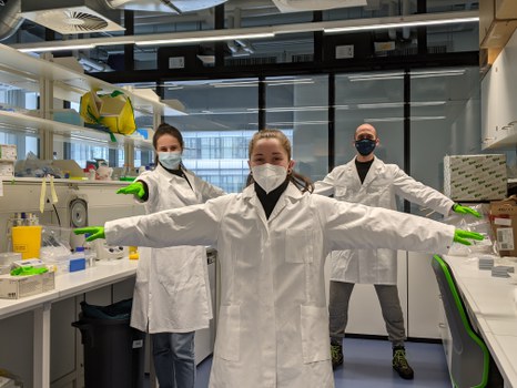 Three young people are standing in a laboratory. They are wearing lab coats and protective masks.