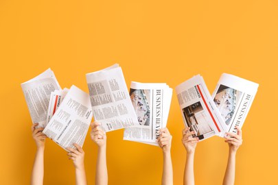 Vier Hände halten verschiedene Zeitungen vor einem leuchtend orangefarbenen Hintergrund hoch. Die Zeitungen zeigen Text und Bilder zu Wirtschaft und Finanzen.