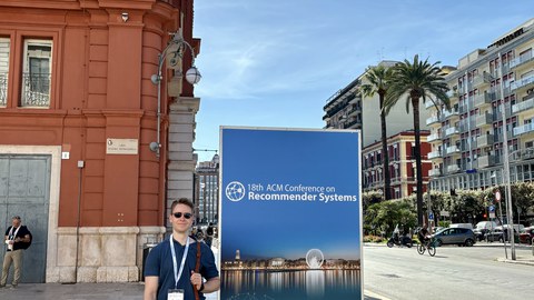 Person standing in front of a large poster for the "18th ACM Conference on Recommender Systems" in Bari, Italy. The poster displays the conference date (October 14-18, 2024) and includes the event logo. In the background, an orange facade and a street wit