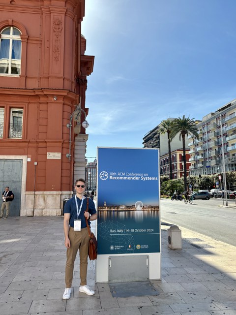 Person standing in front of a large poster for the "18th ACM Conference on Recommender Systems" in Bari, Italy. The poster displays the conference date (October 14-18, 2024) and includes the event logo. In the background, an orange facade and a street wit
