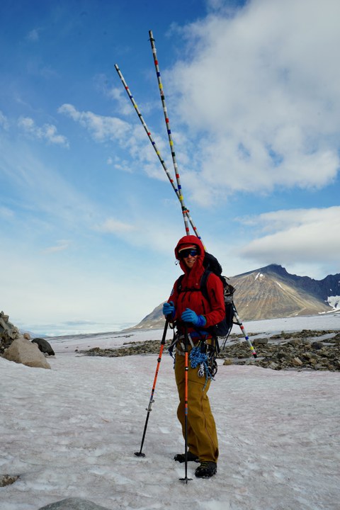 Eine Person in Outdoor-Bekleidung steht in einer eisigen, gebirgigen Landschaft. Sie trägt eine rote Jacke, gelbe Hosen, blaue Handschuhe und eine Sonnenbrille. Auf dem Rücken trägt sie einen Rucksack mit langen, bunten Stangen.
