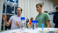 Klemens Bremmel erklärt einem Besucher sein Poster. Im Vordergrund sind verschiedene Flaschen zu sehen. 