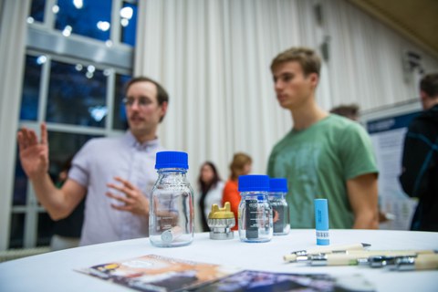 Klemens Bremmel erklärt einem Besucher sein Poster. Im Vordergrund sind verschiedene Flaschen zu sehen. 