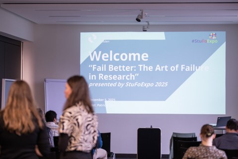 Das Bild zeigt einen Seminarraum und vorne ist eine Präsentation mit einer Begrüßungsfolie zur Veranstaltung zu sehen.