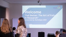 Das Bild zeigt einen Seminarraum und vorne ist eine Präsentation mit einer Begrüßungsfolie zur Veranstaltung zu sehen.