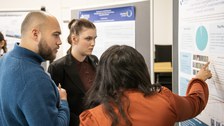 3 Studierende stehen vor einem wissenschaftlichen Poster, das auf einer Pinnwand aufgehängt ist. Eine Studentin zeigt mit dem Finger auf ein Diagramm.