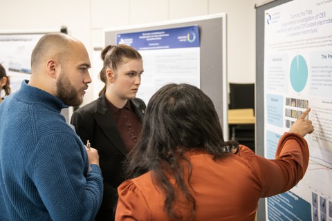 3 Studierende stehen vor einem wissenschaftlichen Poster, das auf einer Pinnwand aufgehängt ist. Eine Studentin zeigt mit dem Finger auf ein Diagramm.