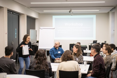 Eine Frau steht vor einer Gruppe Studierender und erklärt etwas. Die Studierenden sitzen an Tischen im Halbkreis darum.