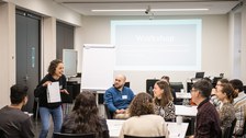Eine Frau steht vor einer Gruppe Studierender und erklärt etwas. Die Studierenden sitzen an Tischen im Halbkreis darum.