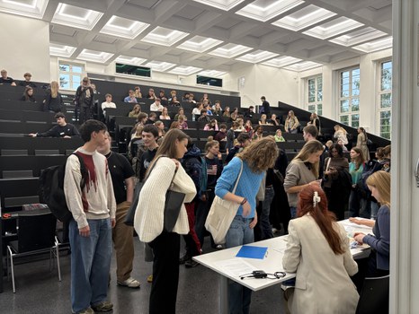 Studierende versammeln sich in einem Hörsaal der TU Dresden. Im Vordergrund melden sich einige Personen an einem Tisch bei zwei Mitarbeiterinnen an, während im Hintergrund viele weitere Studierende in den Sitzreihen Platz nehmen.