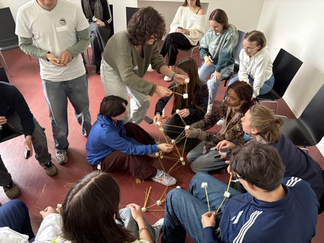 Studierende bauen gemeinsam einen Turm aus Spaghetti und Marshmellows. Einige sitzen, andere stehen und bauen aktiv mit.