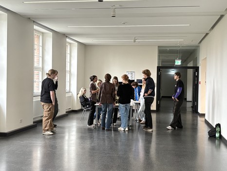 Studierende stehen gemeinsam in einem Flur eines Gebäudes um einen Tisch herum. 