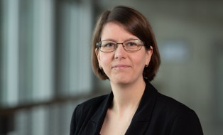 Eine Frau mit kinnlangen braunen Haaren lächelt in die Kamera. Sie hat einen schwarzen Blazer an und trägt eine Brille. 