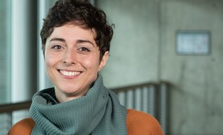 Eine Frau mit kurzen braunen Haaren lächelt in die Kamera. Sie trägt einen orangenen Pullover und einen Schal.