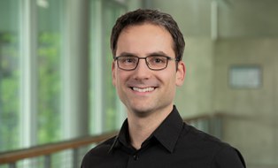Ein Mann mit braunen kurzen Haaren lächelt in die Kamera. Er trägt ein schwarzes Hemd und eine schwarze Brille. 