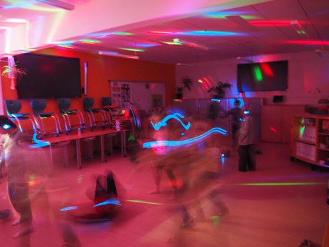 Klassenzimmer bunt beleuchtet bei Nacht, während Kinder tanzen. DIe Kinder sind durch die Bewegung sehr unscharf zu sehen. 