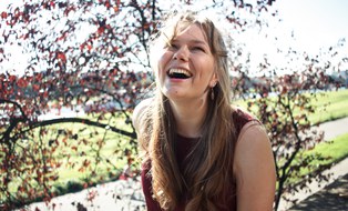 Pia sitzt auf einer Mauer. Hinter ihr befindet sich ein Strauch mit dunkelroten Blättern und eine grüne Wiese. Die Sonne scheint und Pia lacht in die Kamera.