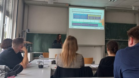 Foto: In einem Seminarraum steht eine Frau vor einer Tafel. Zu sehen sind zudem mehrere Zuhörer:innen in Rückenansicht.